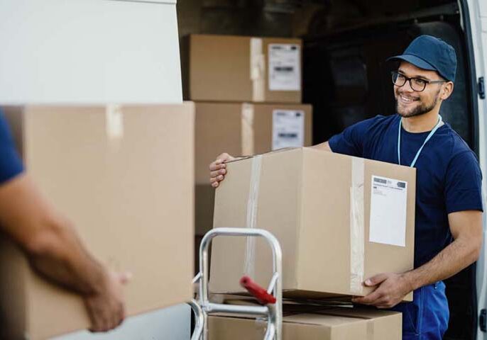 Happy manual worker unloading cardboard boxes from delivery van. Young happy delivery man unloading boxes from a mini van and talking with his coworker.